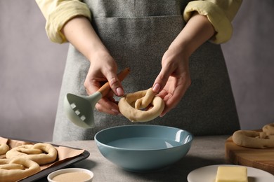 Photo of Woman putting uncooked pretzel into bowl of water at grey table, closeup