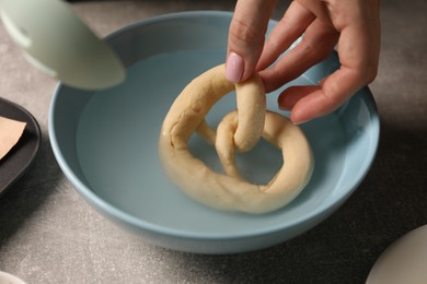 Photo of Woman putting uncooked pretzel into bowl of water at grey table, closeup
