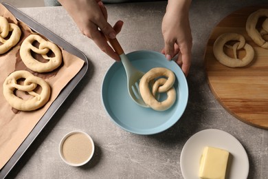 Photo of Woman putting uncooked pretzel into bowl of water at grey table, top view