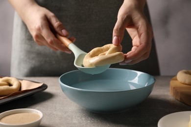 Photo of Woman putting uncooked pretzel into bowl of water at grey table, closeup