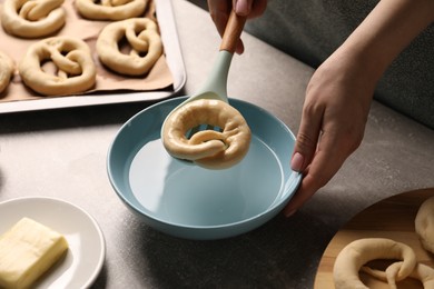 Photo of Woman putting uncooked pretzel into bowl of water at grey table, closeup