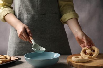 Photo of Woman putting uncooked pretzel into bowl of water at grey table, closeup