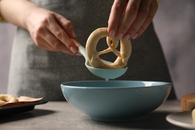 Photo of Woman putting uncooked pretzel into bowl of water at grey table, closeup