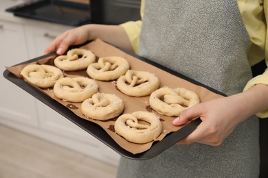 Photo of Woman holding baking dish with uncooked pretzels in kitchen, closeup