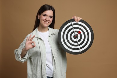 Photo of Targeting. Smiling woman with dart board and arrow showing ok gesture on beige background
