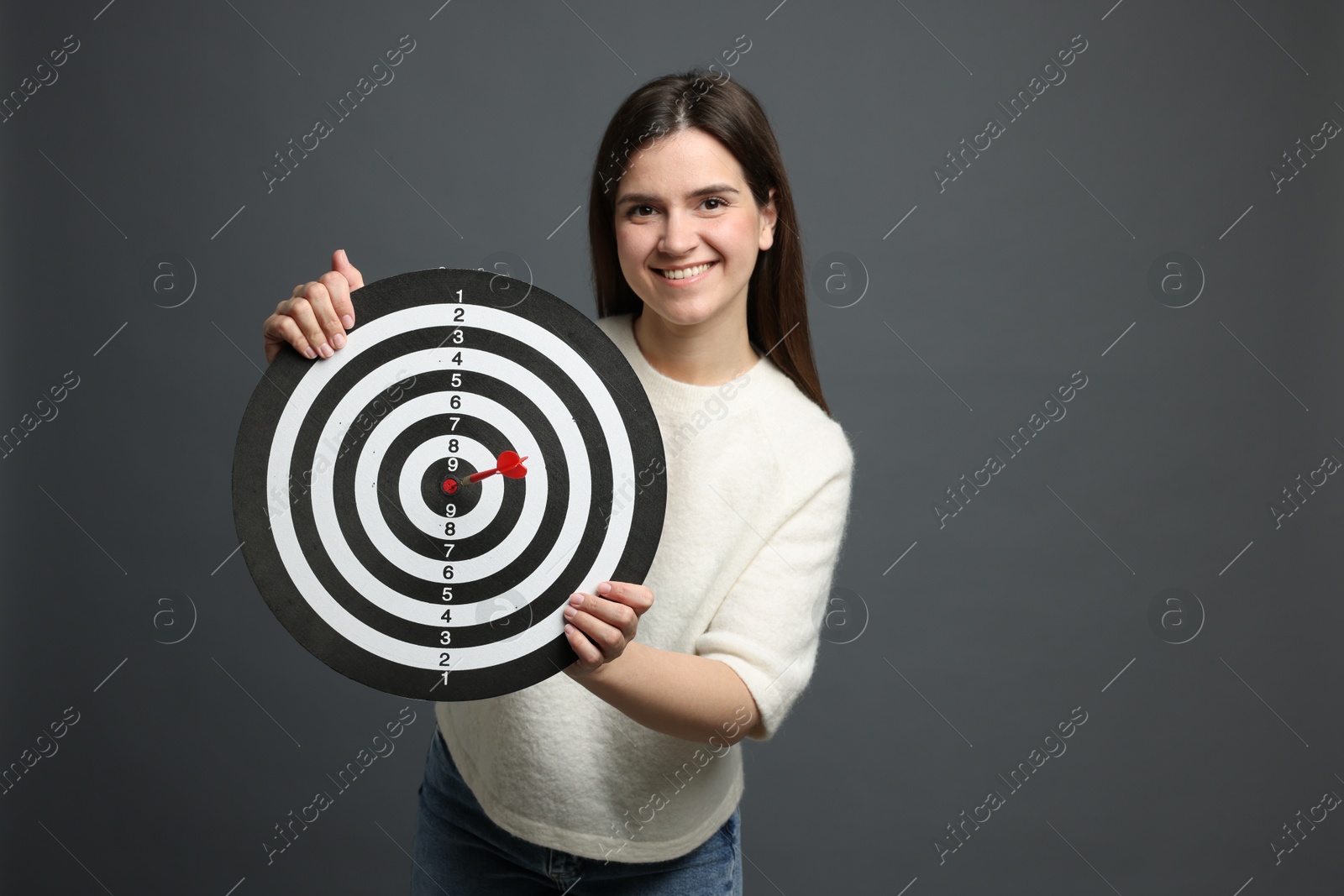 Targeting. Smiling woman with dart board and arrow on grey background Photo of Targeting. Smiling woman with dart board and arrow on grey background