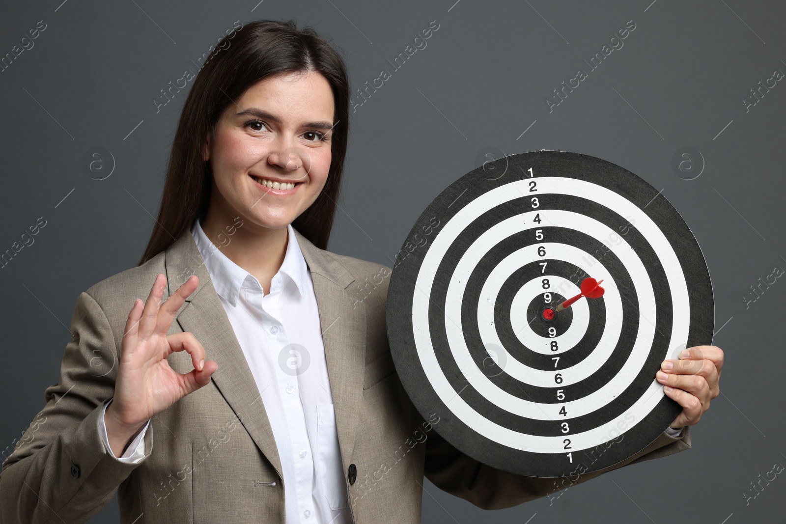 Targeting. Smiling businesswoman with dart board and arrow showing ok gesture on grey background Photo of Targeting. Smiling businesswoman with dart board and arrow showing ok gesture on grey background