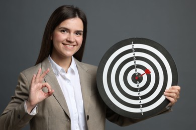 Photo of Targeting. Smiling businesswoman with dart board and arrow showing ok gesture on grey background