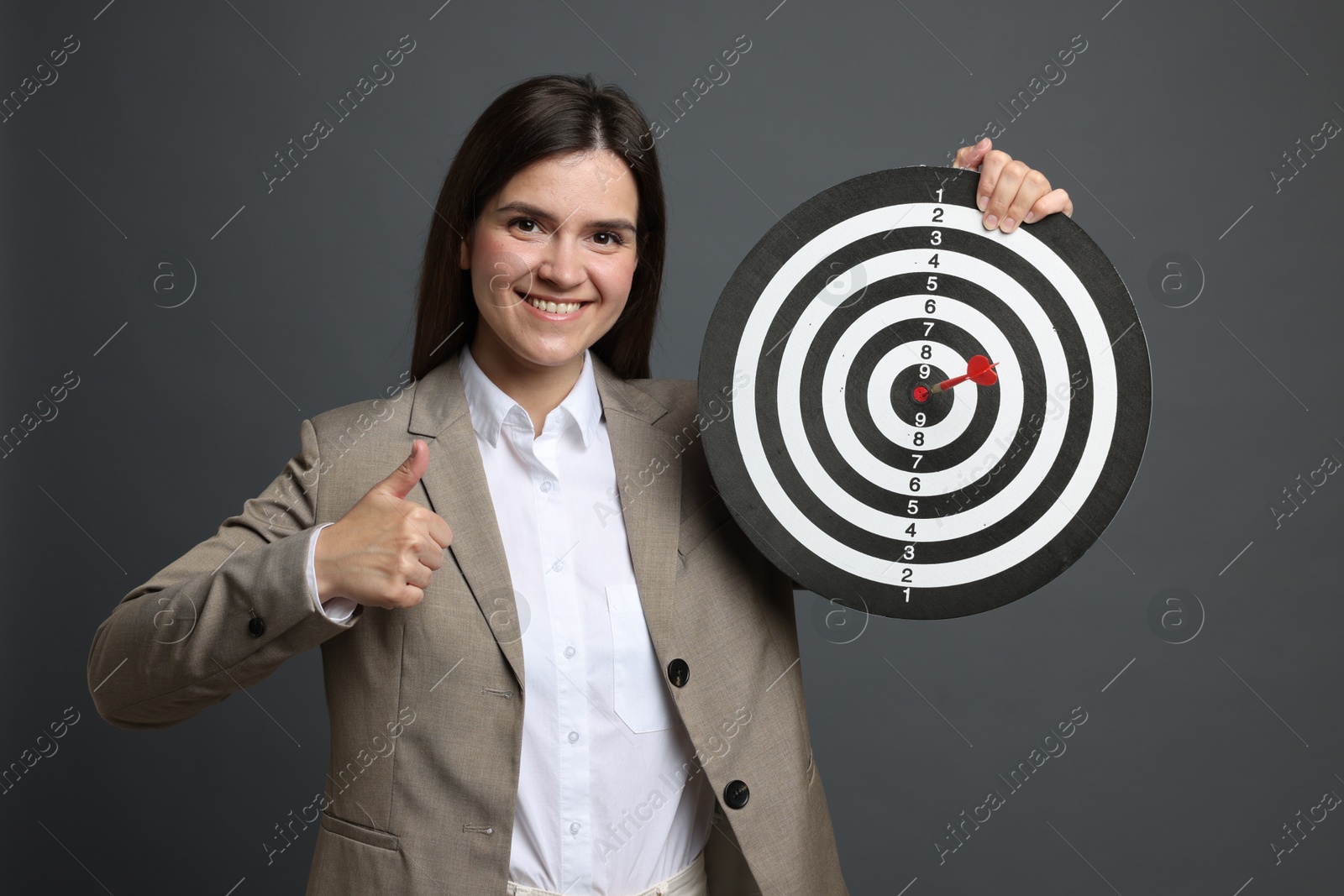 Targeting. Smiling businesswoman with dart board and arrow showing thumbs up on grey background Photo of Targeting. Smiling businesswoman with dart board and arrow showing thumbs up on grey background