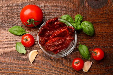 Photo of Delicious sun dried tomatoes in glass bowl, fresh vegetables and spices on wooden table, flat lay