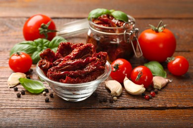 Photo of Delicious sun dried tomatoes in glass dishware, fresh vegetables and spices on wooden table, closeup