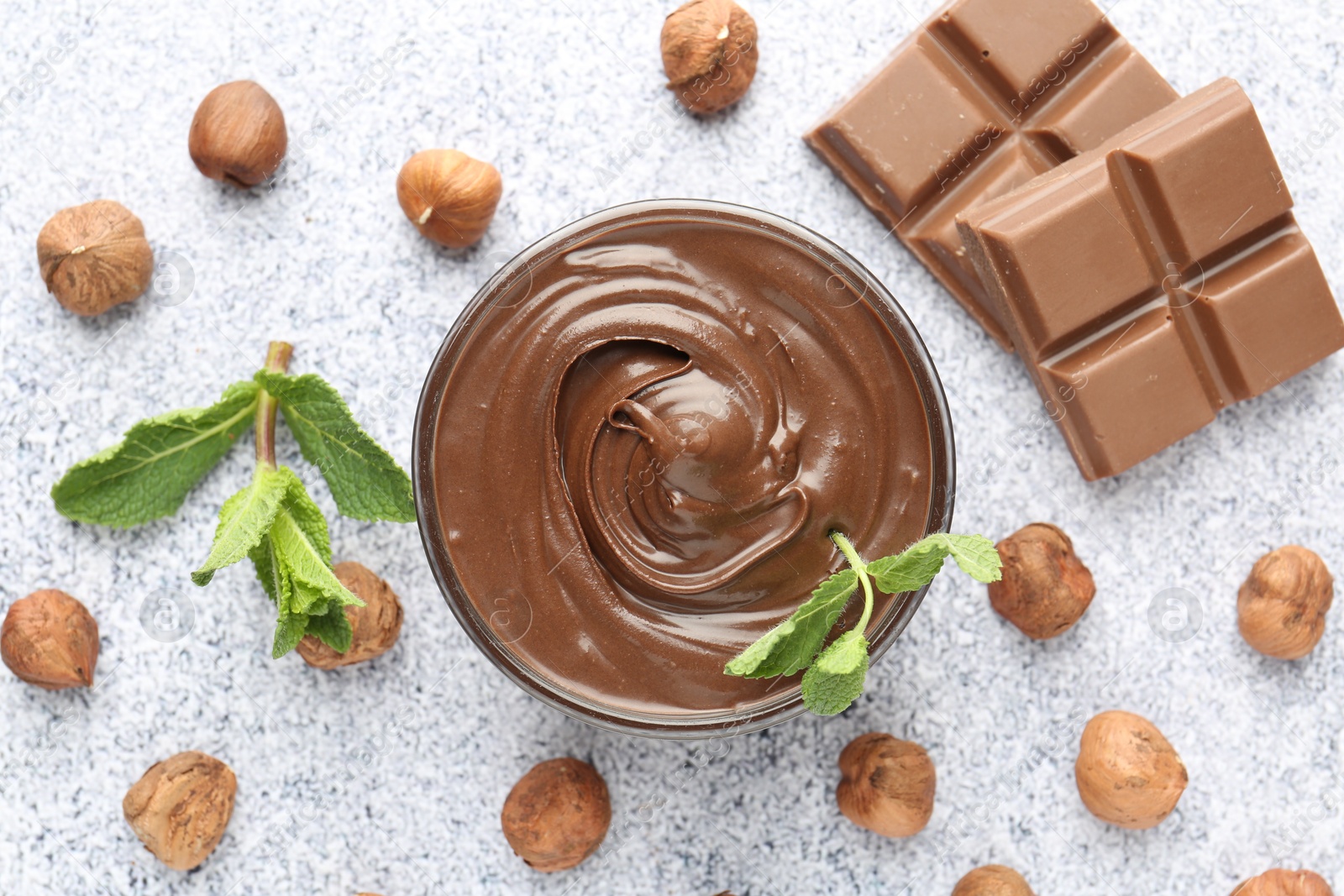 Photo of Tasty chocolate spread with mint in bowl and hazelnuts on grey textured table, flat lay
