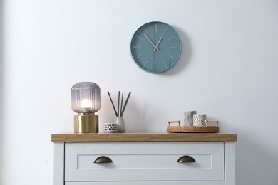 Photo of Dresser with stylish reed diffuser and lamp near white wall indoors