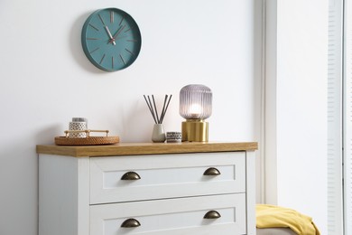 Photo of Dresser with stylish reed diffuser and lamp near white wall indoors