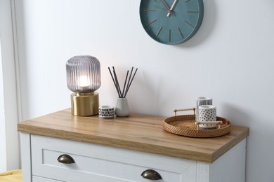 Photo of Dresser with stylish reed diffuser and lamp near white wall indoors