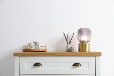 Photo of Dresser with stylish reed diffuser and lamp near white wall indoors