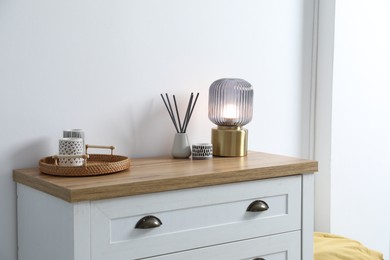 Photo of Dresser with stylish reed diffuser and lamp near white wall indoors