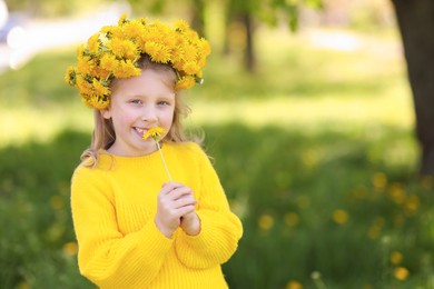 Photo of Cute little girl with beautiful flower wearing wreath of dandelions in park, space for text