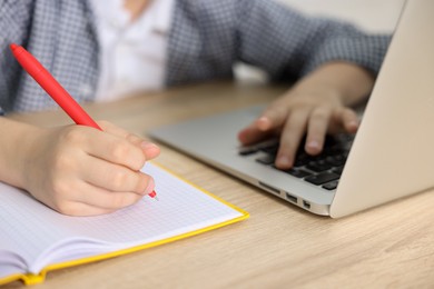 Photo of Little boy doing homework with laptop at wooden table indoors, closeup
