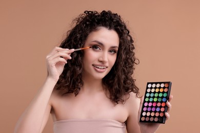Photo of Smiling woman with eyeshadow palette and brush applying makeup on beige background