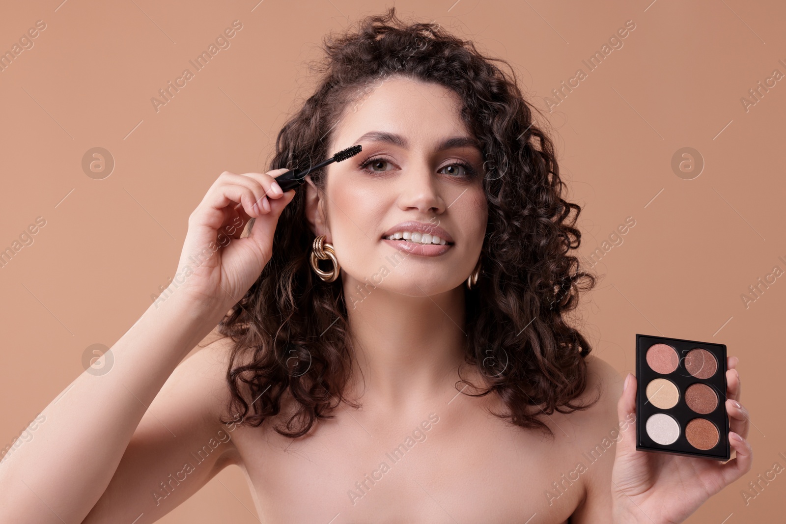 Smiling woman with eyeshadow palette and brush applying makeup on beige background Photo of Smiling woman with eyeshadow palette and brush applying makeup on beige background