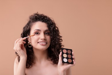 Photo of Smiling woman with eyeshadow palette and brush applying makeup on beige background. Space for text