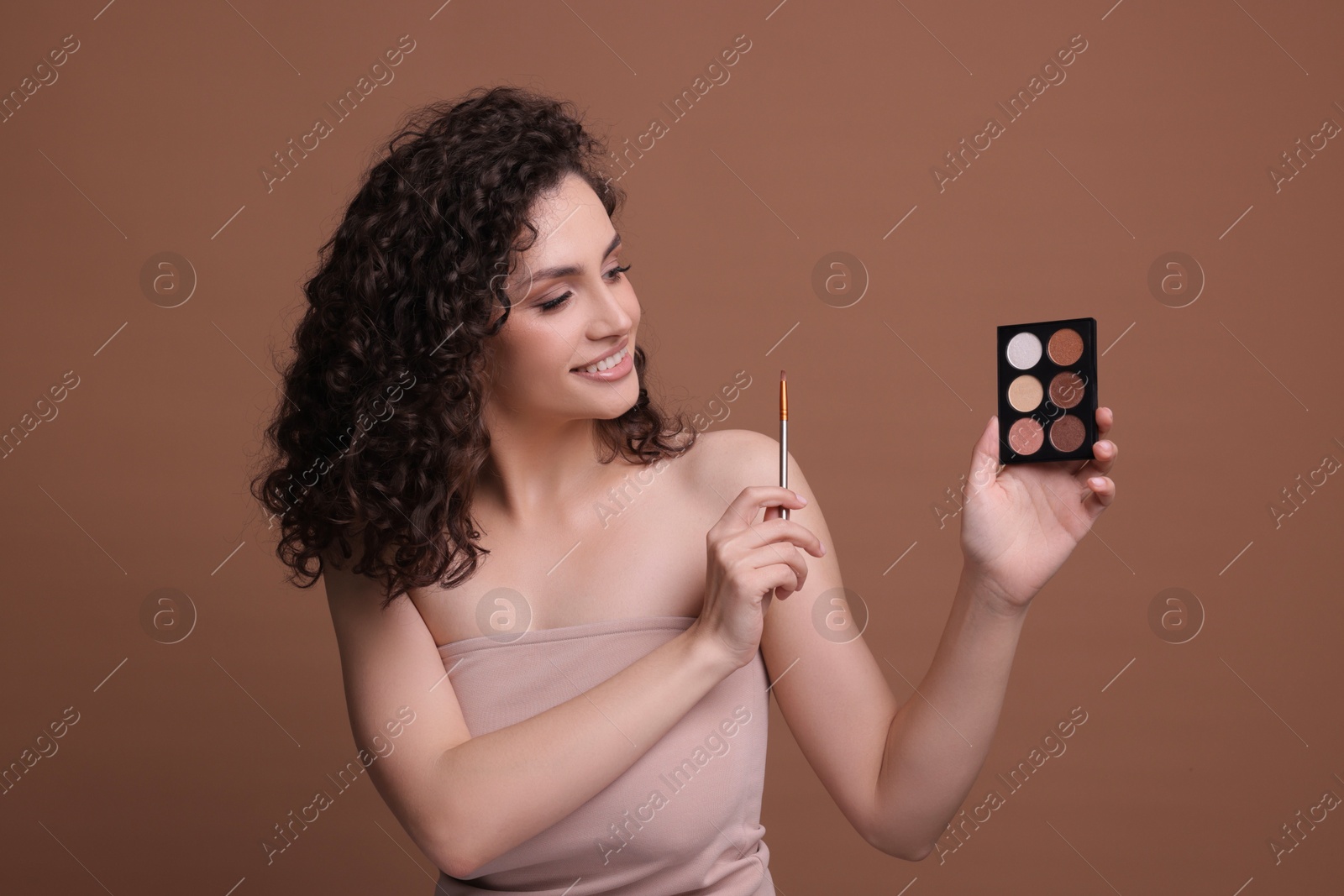 Smiling woman with eyeshadow palette and brush on brown background Photo of Smiling woman with eyeshadow palette and brush on brown background