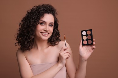 Photo of Smiling woman with eyeshadow palette and brush on brown background
