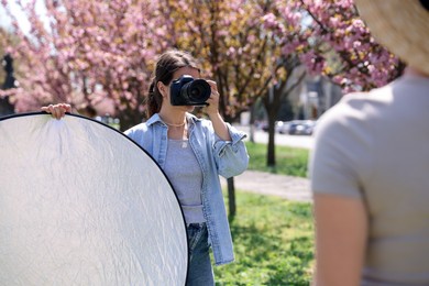 Photo of Professional photographer with camera and reflector near model during shooting outdoors on sunny day