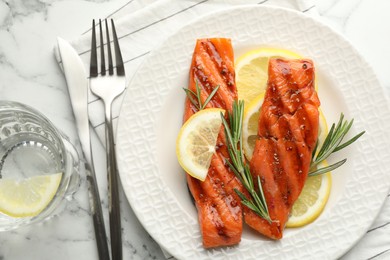 Photo of Seafood. Tasty grilled salmon served with water, cutlery, rosemary and lemon on white marble table, flat lay