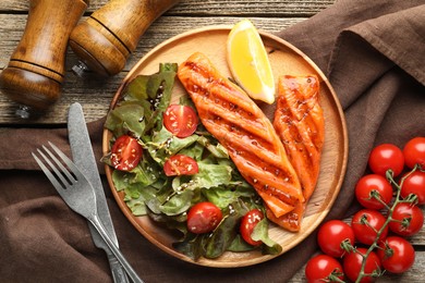 Photo of Tasty grilled salmon served with cutlery, salad, tomatoes and lemon on wooden table, flat lay