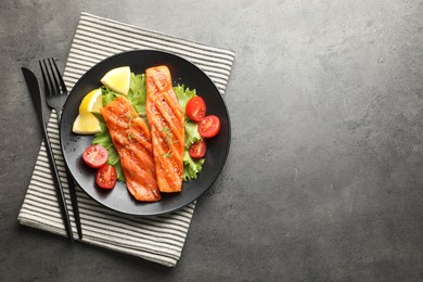 Photo of Tasty grilled salmon served with cutlery, lettuce, tomatoes and lemon on grey table, top view. Space for text