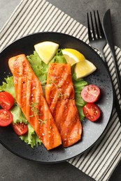 Photo of Tasty grilled salmon served with cutlery, lettuce, tomatoes and lemon on grey table, top view
