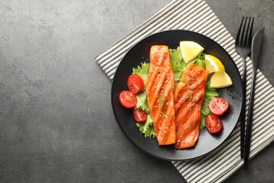 Photo of Tasty grilled salmon served with cutlery, lettuce, tomatoes and lemon on grey table, top view. Space for text