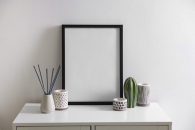 Photo of Empty picture frame, reed diffuser and stylish home decor on cabinet near white wall indoors