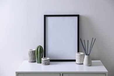 Photo of Empty picture frame, reed diffuser and stylish home decor on cabinet near white wall indoors