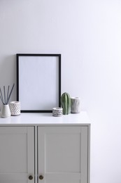 Photo of Empty picture frame, reed diffuser and stylish home decor on cabinet near white wall indoors