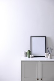 Photo of Empty picture frame, reed diffuser and stylish home decor on cabinet near white wall indoors