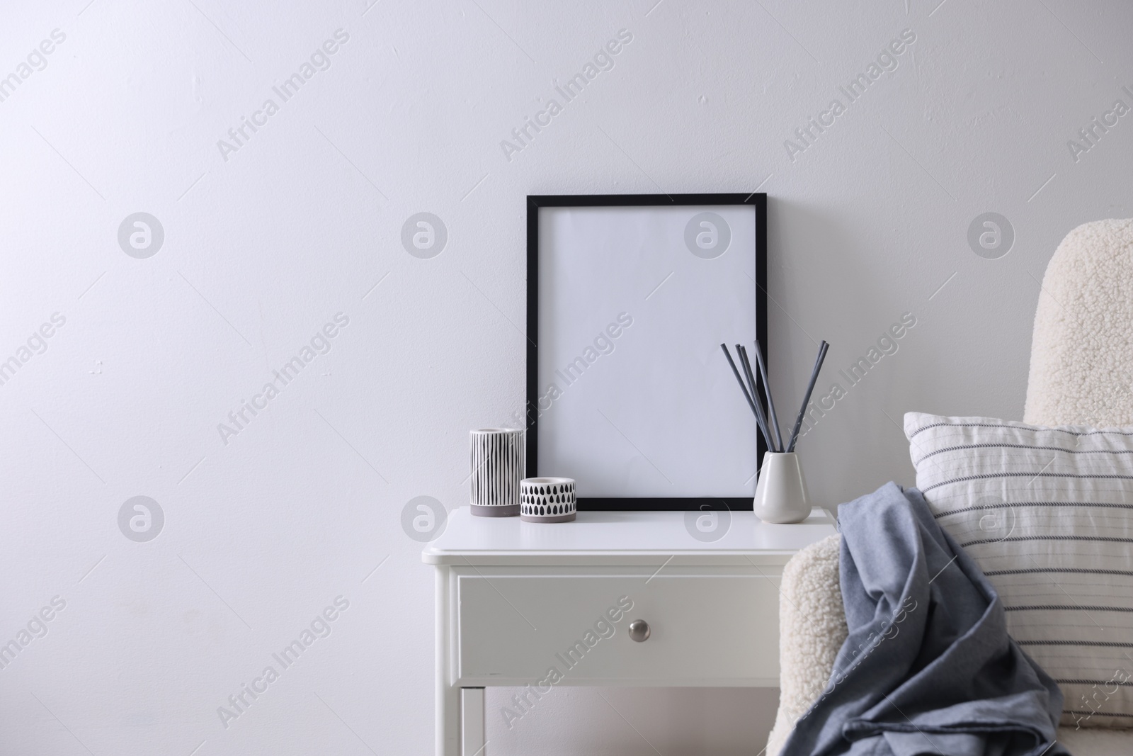 Empty picture frame, reed diffuser, armchair and stylish home decor on table near white wall indoors Photo of Empty picture frame, reed diffuser, armchair and stylish home decor on table near white wall indoors