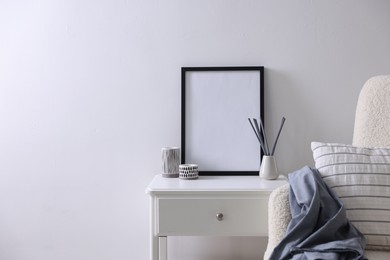 Photo of Empty picture frame, reed diffuser, armchair and stylish home decor on table near white wall indoors