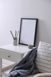 Photo of Empty picture frame, reed diffuser, armchair and stylish home decor on table near white wall indoors