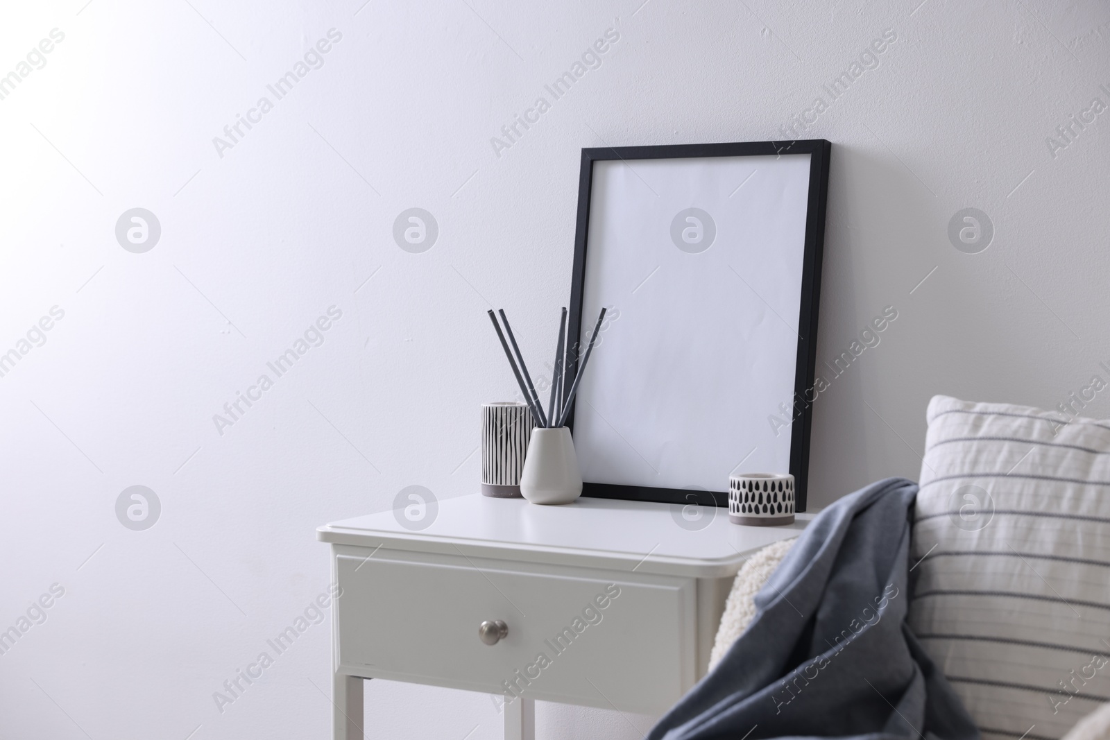 Empty picture frame, reed diffuser, armchair and stylish home decor on table near white wall indoors Photo of Empty picture frame, reed diffuser, armchair and stylish home decor on table near white wall indoors