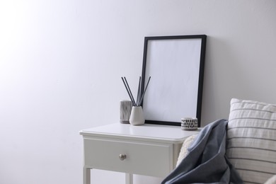 Photo of Empty picture frame, reed diffuser, armchair and stylish home decor on table near white wall indoors