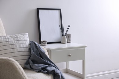 Photo of Empty picture frame, reed diffuser, armchair and stylish home decor on table near white wall indoors