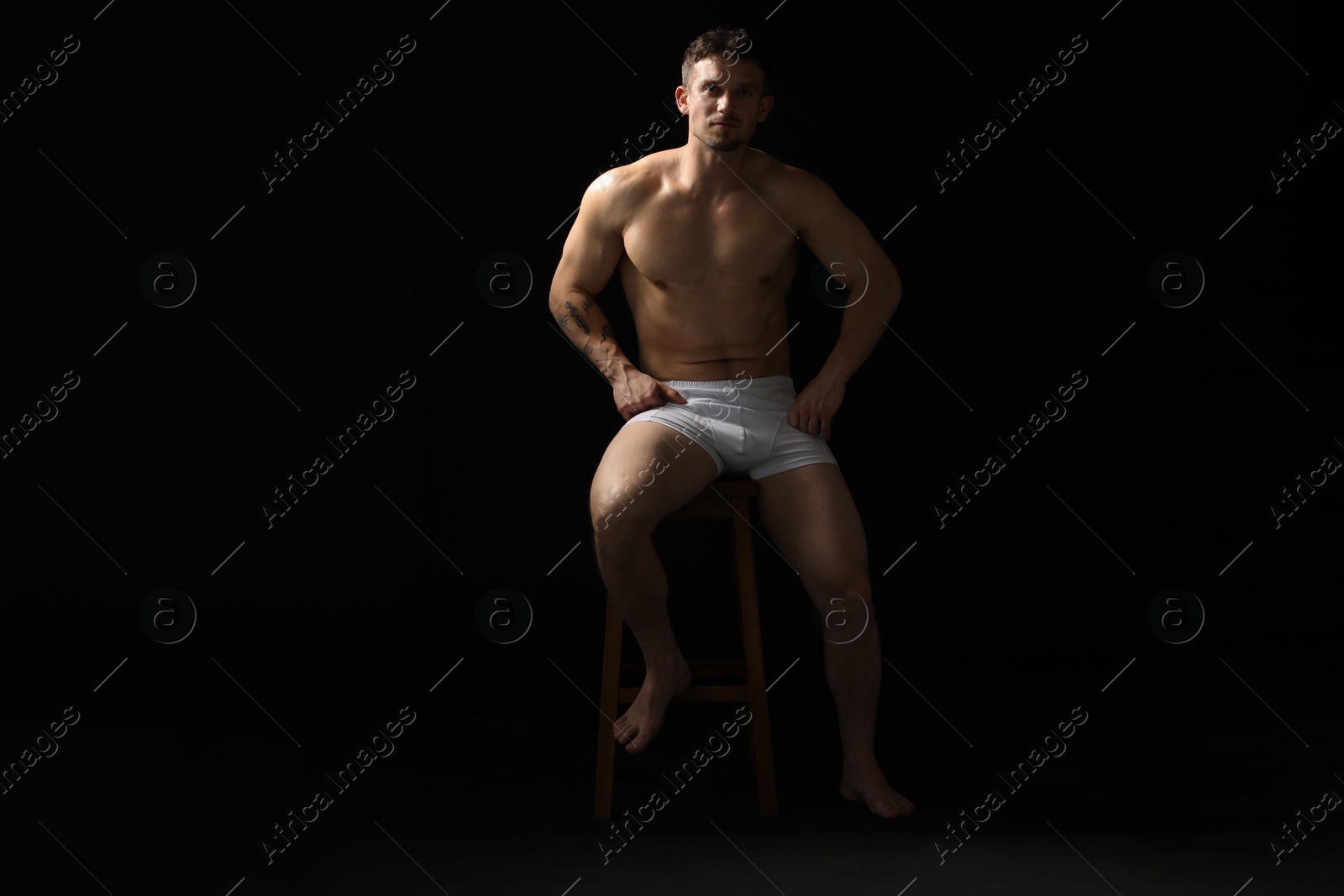 Young man in stylish white underwear on chair against black background Photo of Young man in stylish white underwear on chair against black background