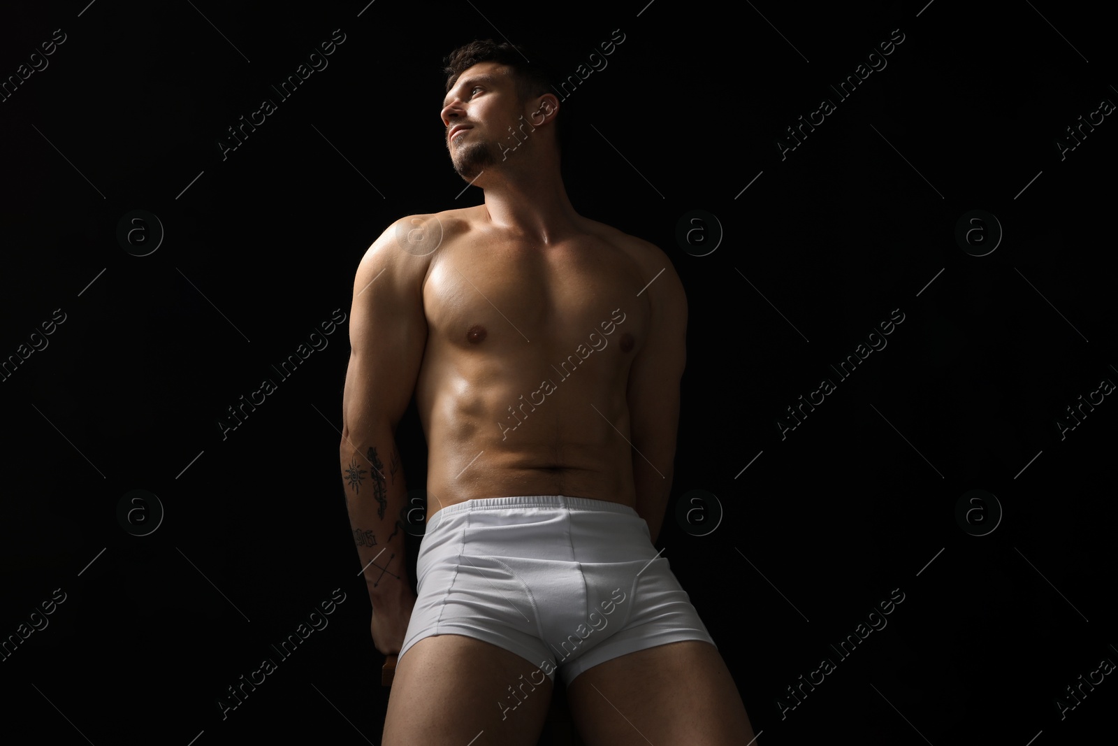 Young man in stylish white underwear on chair against black background, low angle view Photo of Young man in stylish white underwear on chair against black background, low angle view