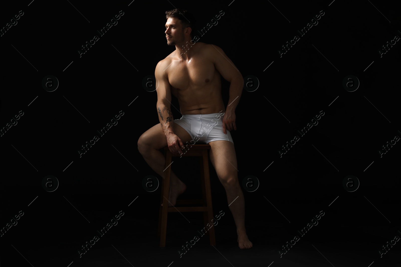 Young man in stylish white underwear on chair against black background Photo of Young man in stylish white underwear on chair against black background