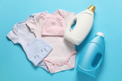 Photo of Bottles of fabric softener and baby clothes on light blue background, flat lay