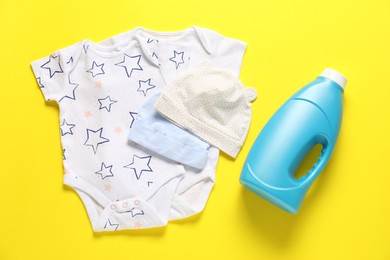 Photo of Bottle of fabric softener and baby clothes on yellow background, flat lay