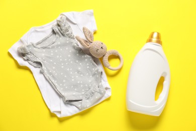 Photo of Bottle of fabric softener, toy and baby clothes on yellow background, flat lay